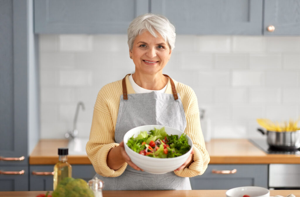 A senior woman in the kitchen holding a bowl of salad on both hands, smiling and looking directly at the camera.